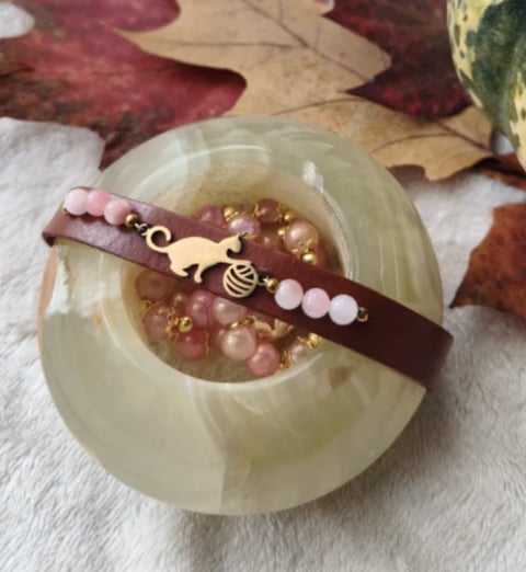 Beaded bracelet with burgundy leather band, rose quartz beads, and gold cat charm displayed on pale jade stone with autumn leaves.